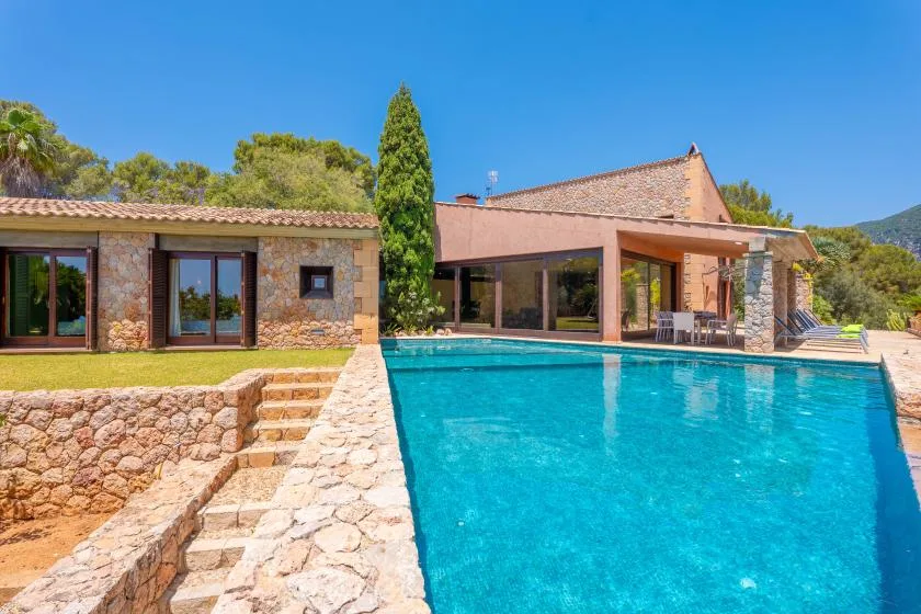 A stone house opens onto a swimming pool and a shaded outdoor seating area.