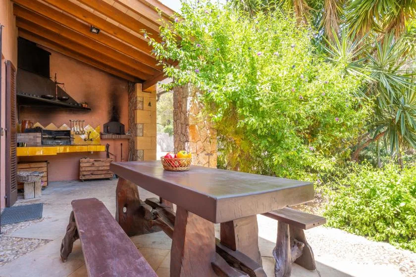 An outdoor dining area with a wooden table beside a covered cooking space and greenery.