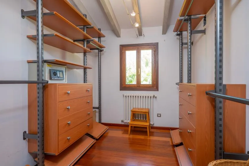 A narrow storage room with wooden shelving, drawer units, and a window above a radiator.