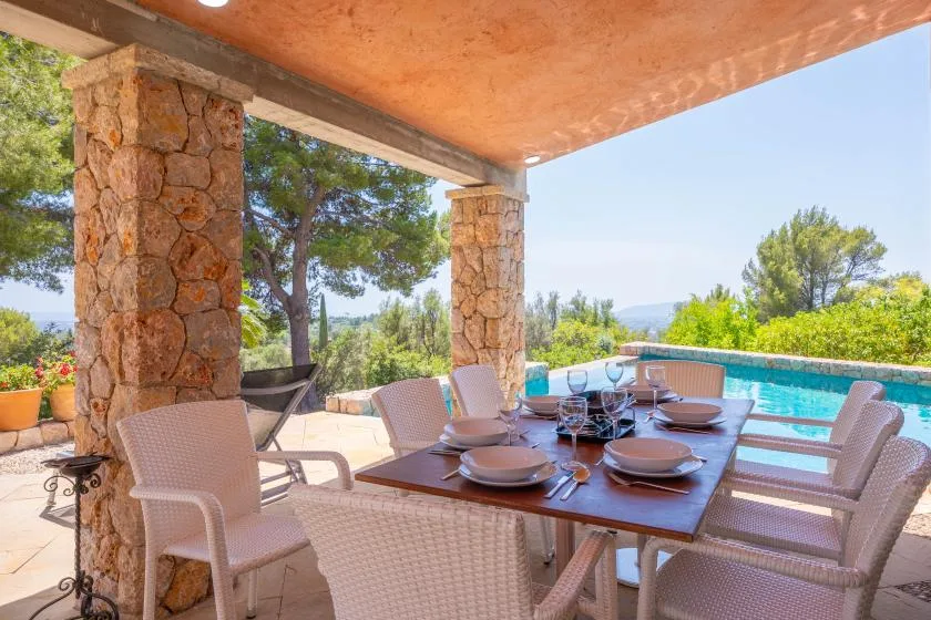 A covered patio dining area overlooks a swimming pool and surrounding trees.