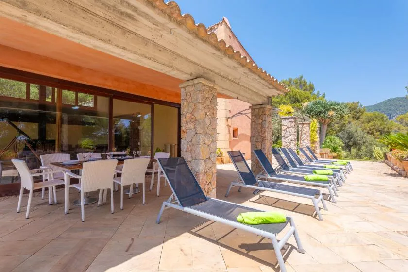A sunny stone patio has outdoor seating beside a row of lounge chairs.