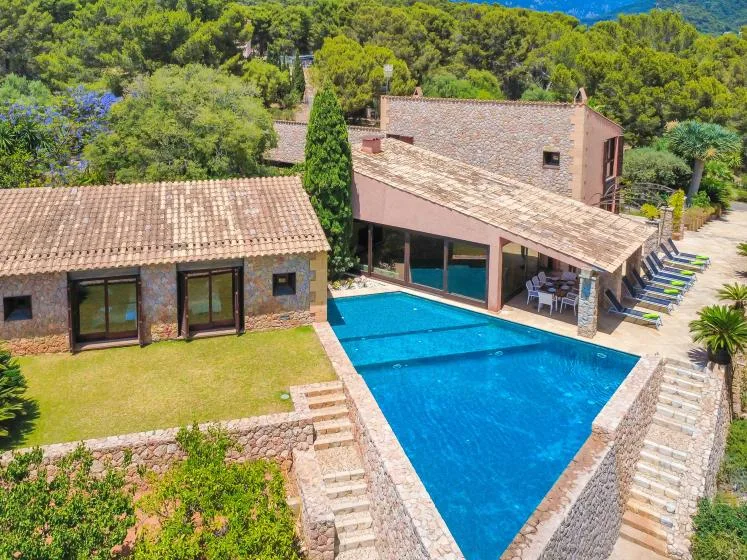 A swimming pool sits beside a stone house and a covered outdoor seating area.