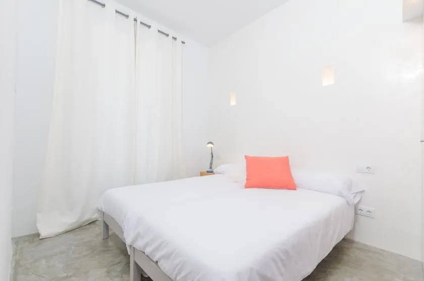 A bright bedroom with a white bed, floor-length curtains, and a coral accent pillow.