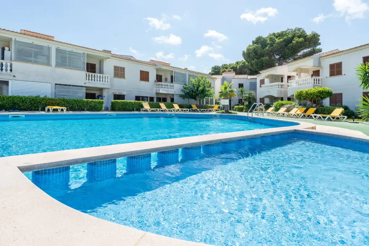 A swimming pool sits in front of white buildings and rows of sun loungers.