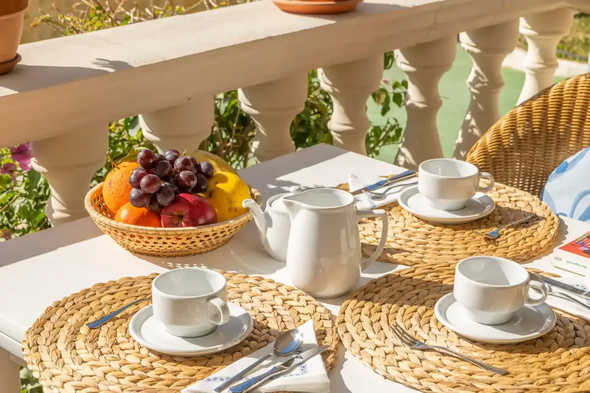 A sunlit outdoor dining table is set with cups and a basket of fruit.