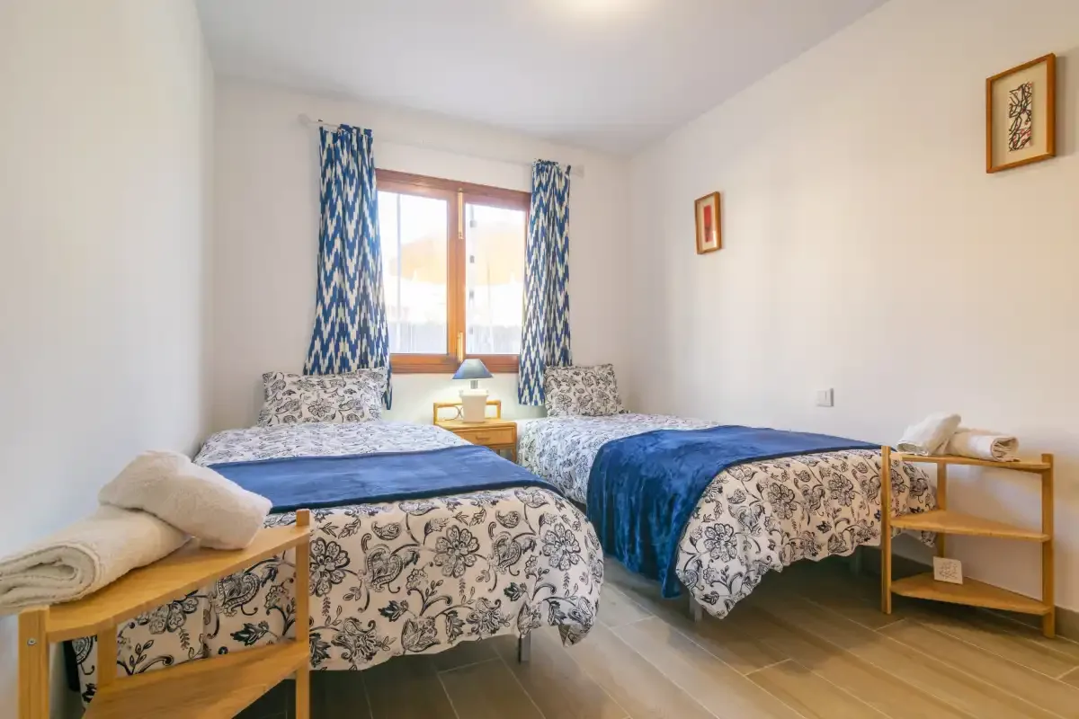 A bright bedroom with two beds, patterned bedding, and blue curtains.