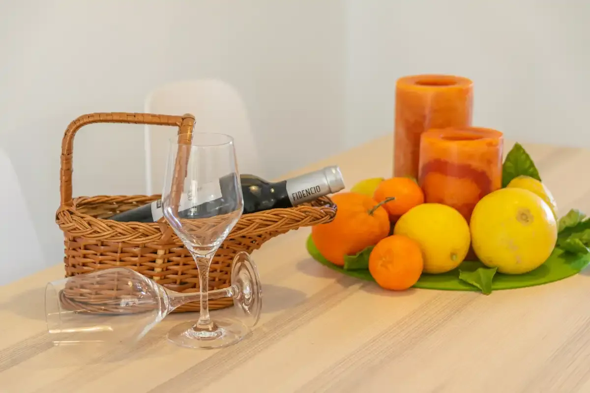 A tabletop displays a wicker basket, wine glasses, oranges, lemons, and candles.