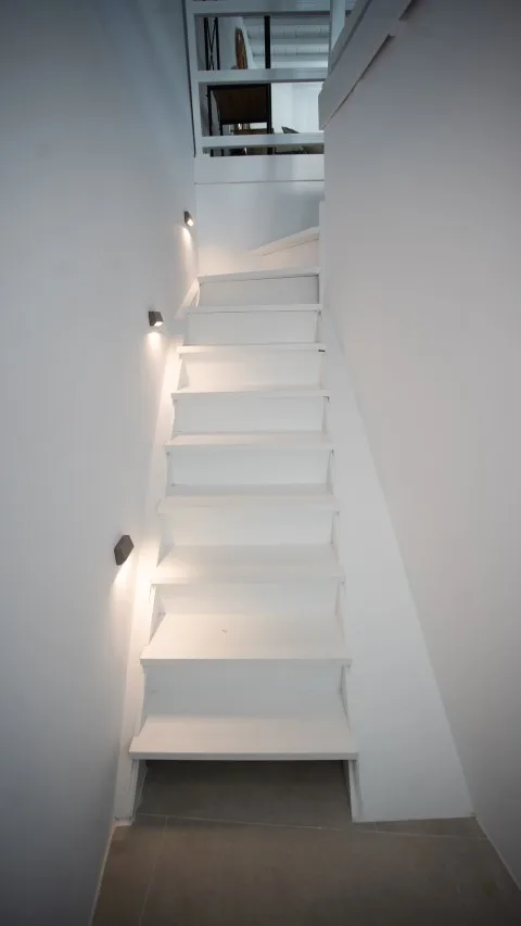 Una estrecha escalera blanca sube hacia una abertura con paneles de vidrio.