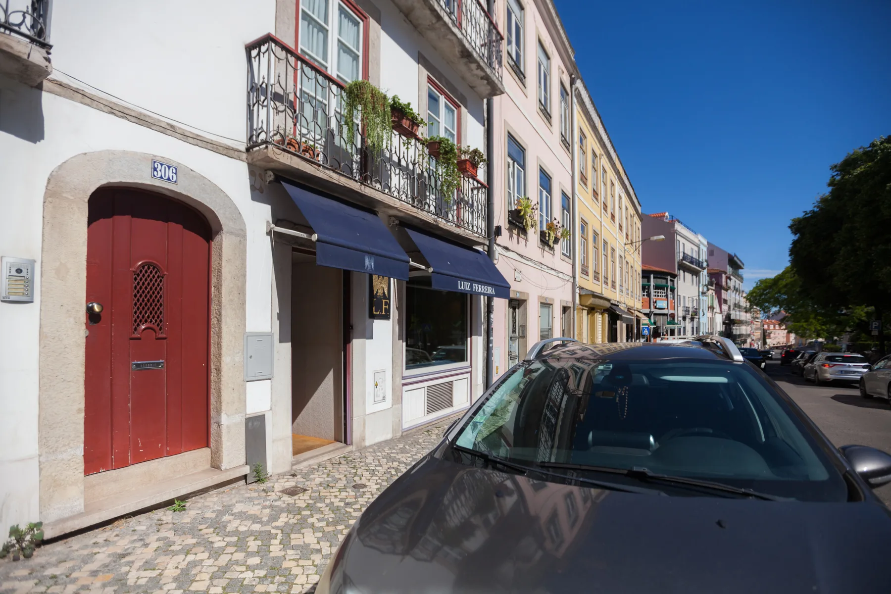Una calle urbana soleada con fachadas coloridas, coches estacionados y una acera empedrada.