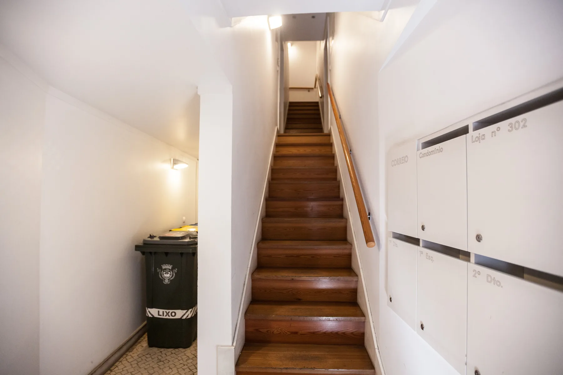 Una escalera interior con peldaños de madera, buzones y un cubo de basura.