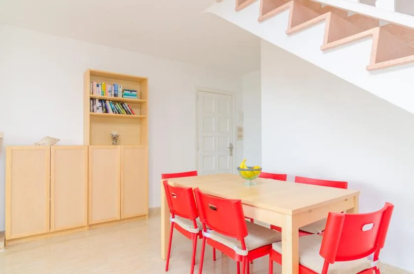 A dining area with a light wood table and red chairs beneath a staircase.