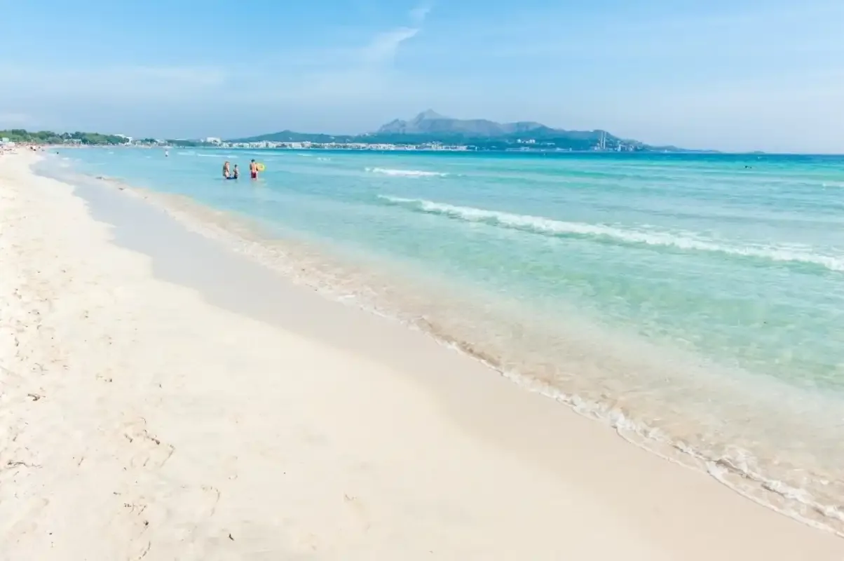 A sandy beach curves beside clear turquoise water, with distant mountains on the horizon.