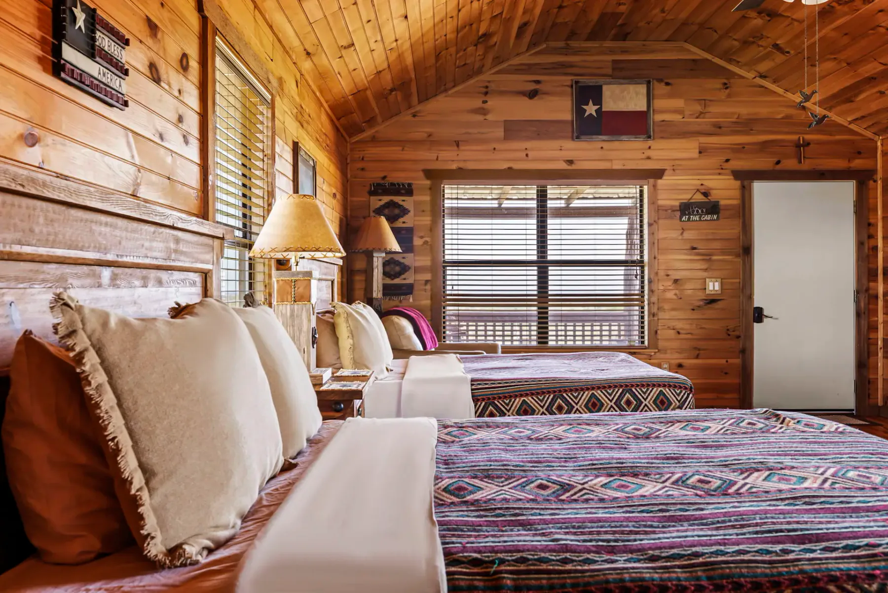 Dormitorio rústico con paredes revestidas de madera, dos camas con colchas estampadas, una lámpara entre ellas y una ventana con persianas.