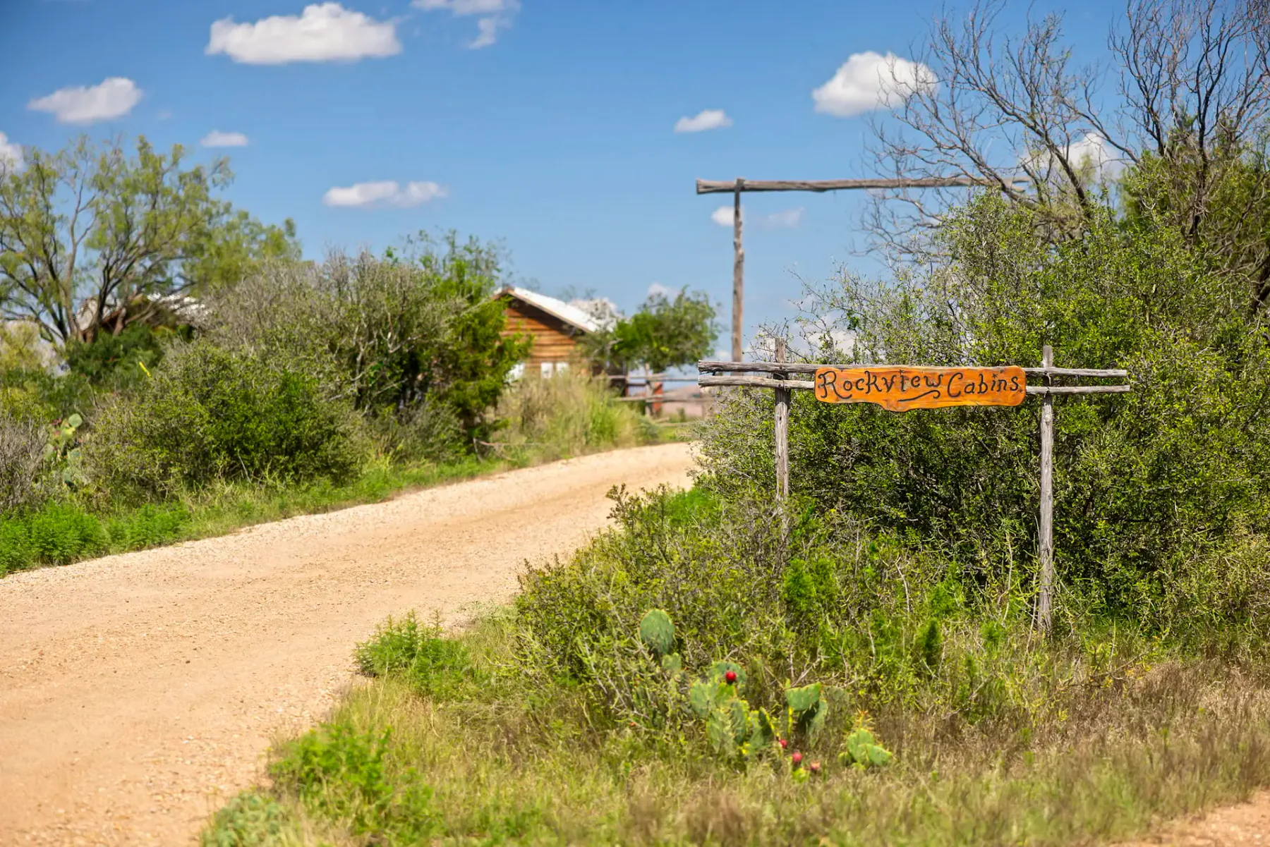 Camino de tierra que conduce a una cabaña rústica, rodeada de vegetación, con un cartel que dice