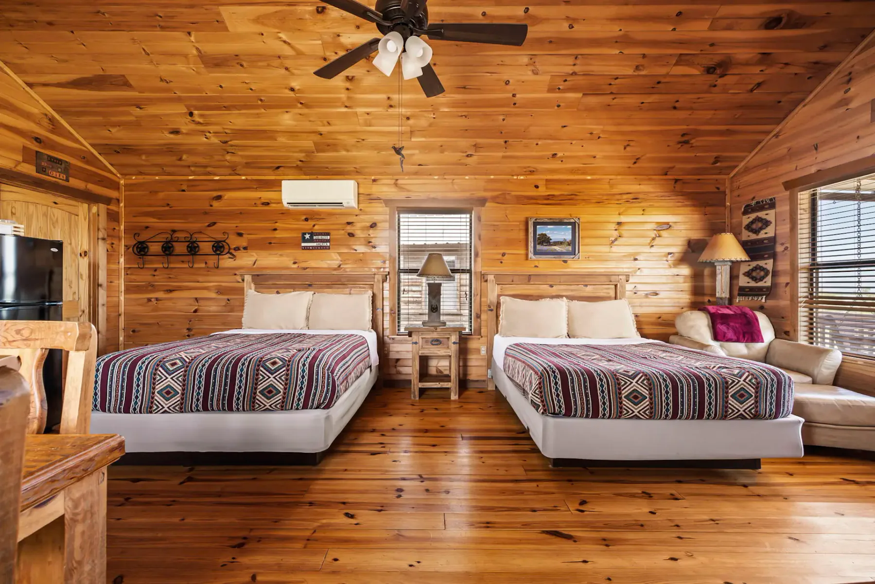 Dormitorio de cabaña rústica con dos camas dobles, paredes de madera, un ventilador de techo y un sofá beige. La decoración incluye cuadros y una pequeña lámpara de mesa.