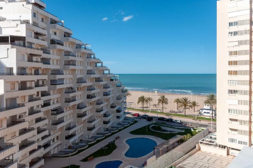 Apartment buildings overlook a beachfront, with a pool and landscaped grounds in the foreground.