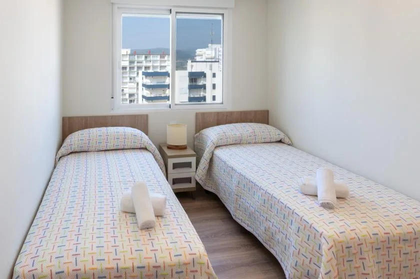 A bright bedroom has two single beds beside a window overlooking nearby buildings.