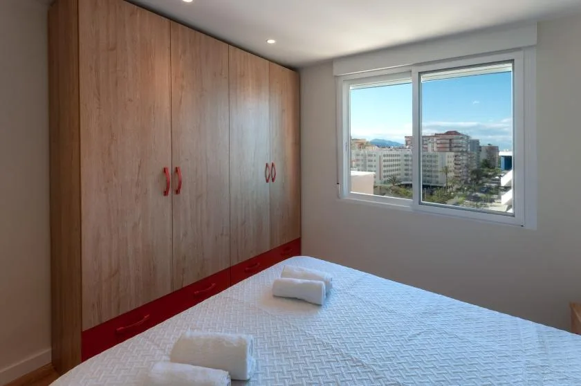 A bedroom with a bed beside a large window overlooking nearby buildings.