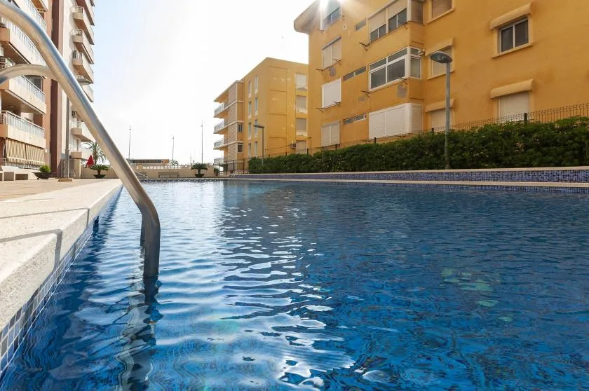 An outdoor swimming pool sits between apartment buildings, with a metal handrail at the edge.