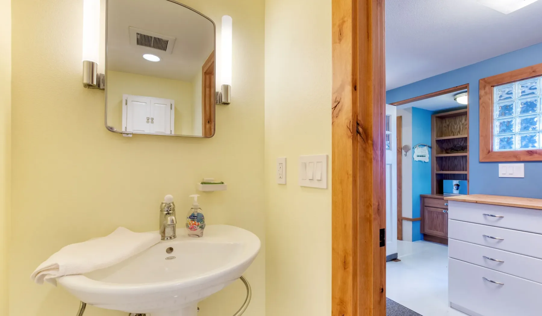 Un lavabo y un espejo de baño dan a una habitación azul con cajones y estantes.