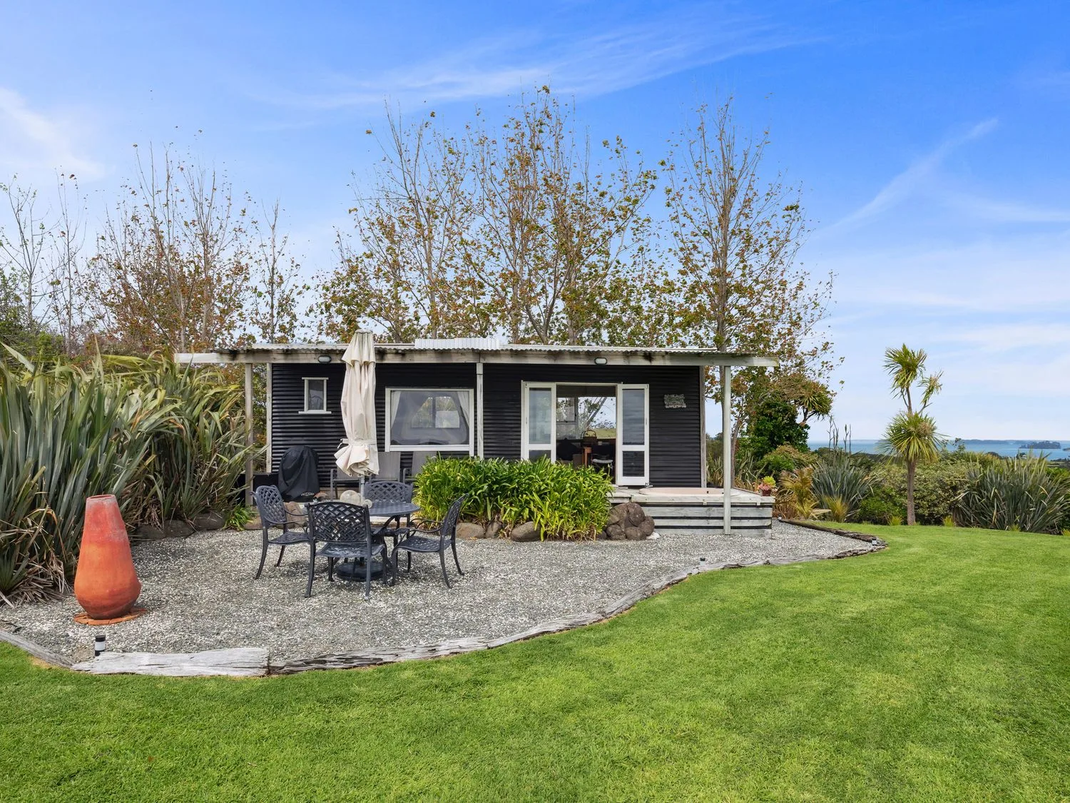 A small black cabin with a white roof surrounded by trees and bushes with a gravel patio and outdoor table with chairs at Tawharanui Cabin - Rural Cabin Studio in Tawharanui