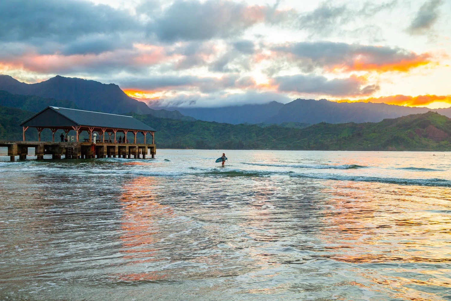 Una persona camina por aguas poco profundas bajo un atardecer colorido y montañas lejanas.