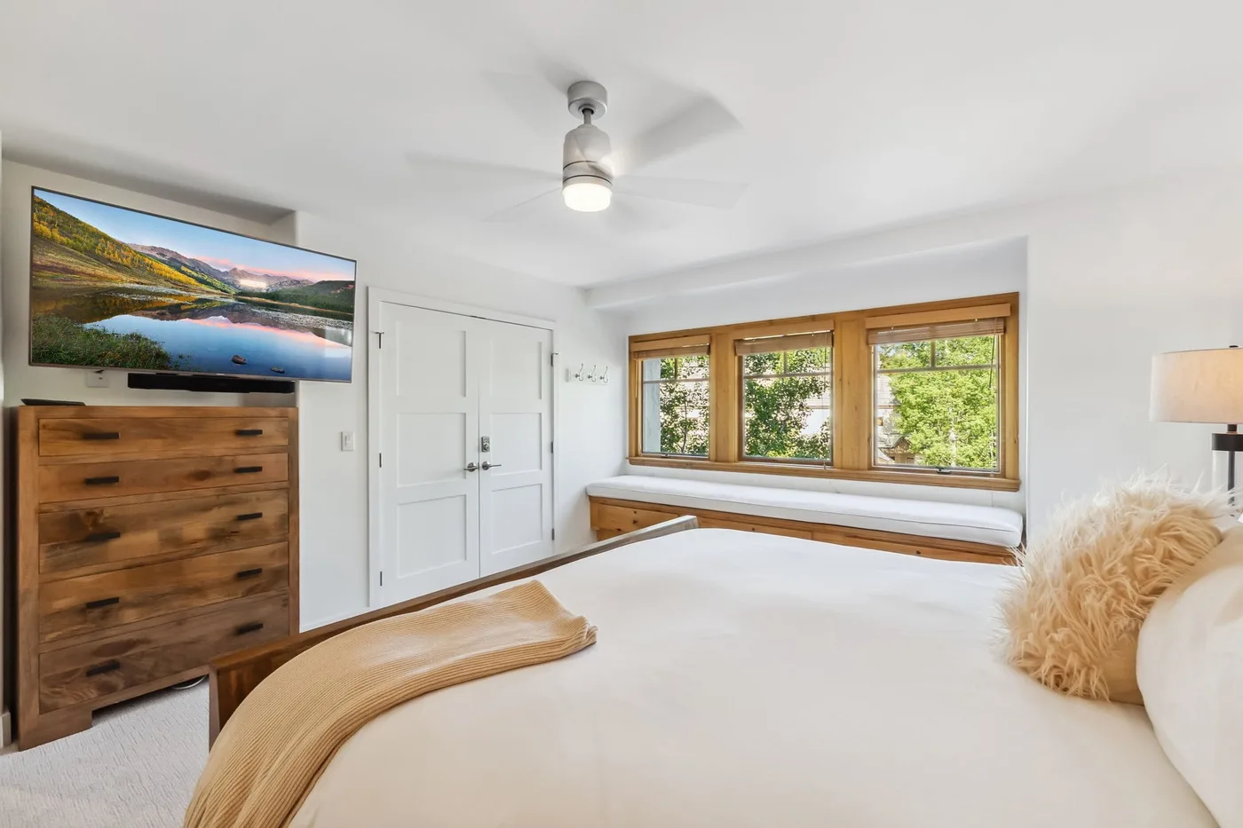 Un dormitorio luminoso con una cama, una cómoda de madera y ventanas con vistas a la vegetación.