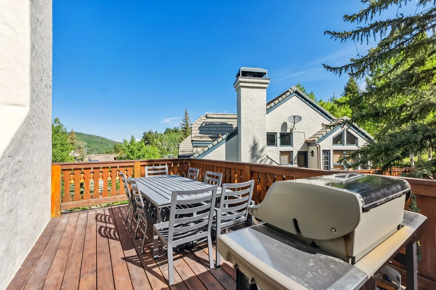 Una terraza de madera con asientos al aire libre y una parrilla grande da a casas y árboles cercanos.