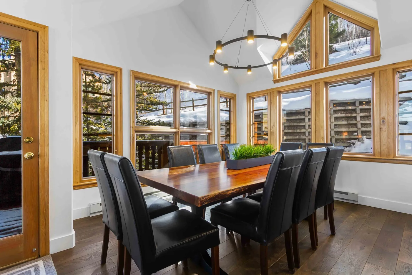 A dining area with a long wooden table and dark seating beside large windows.