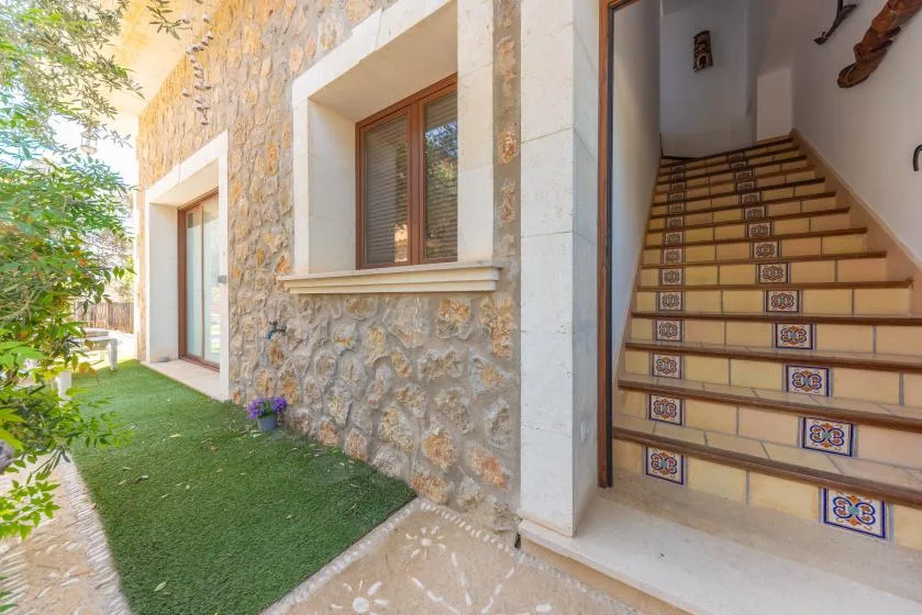 Una pared exterior de piedra bordea un estrecho paso con césped junto a una escalera de azulejos.