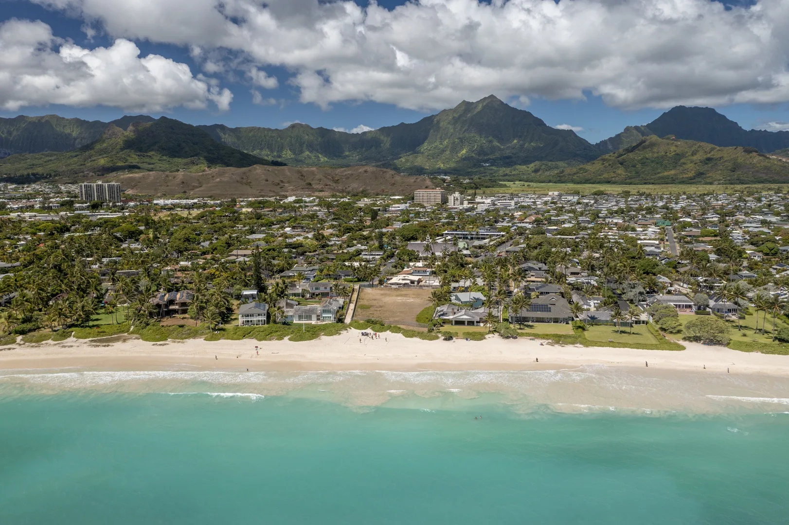 An aerial view shows a sandy beach, turquoise water, and a town beneath green mountains.