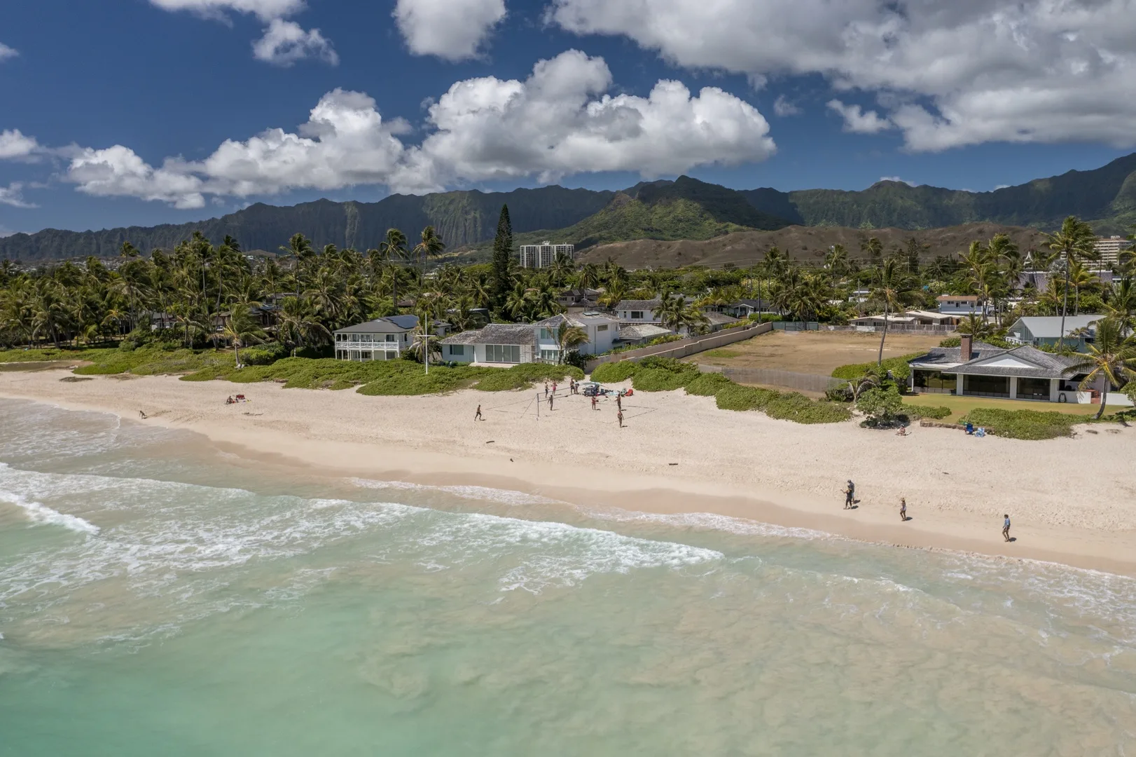 A sandy beach borders turquoise water, with homes and green mountains beyond.