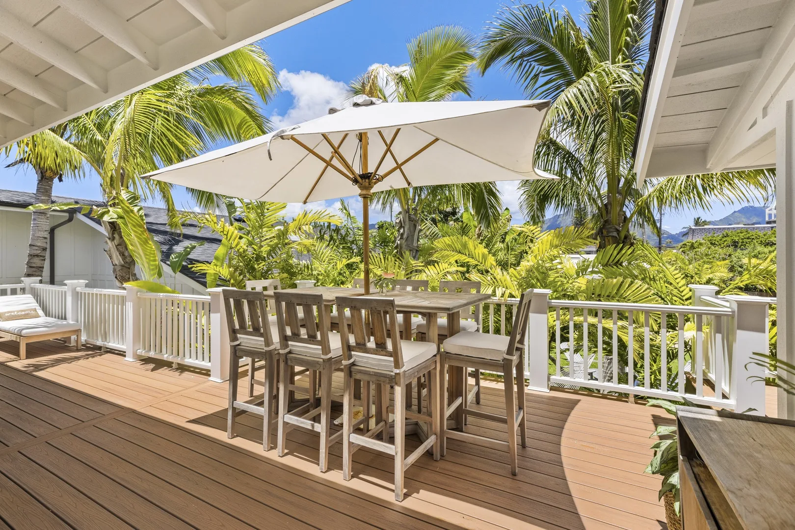 An outdoor deck has dining seating beneath a large umbrella, surrounded by palm trees.