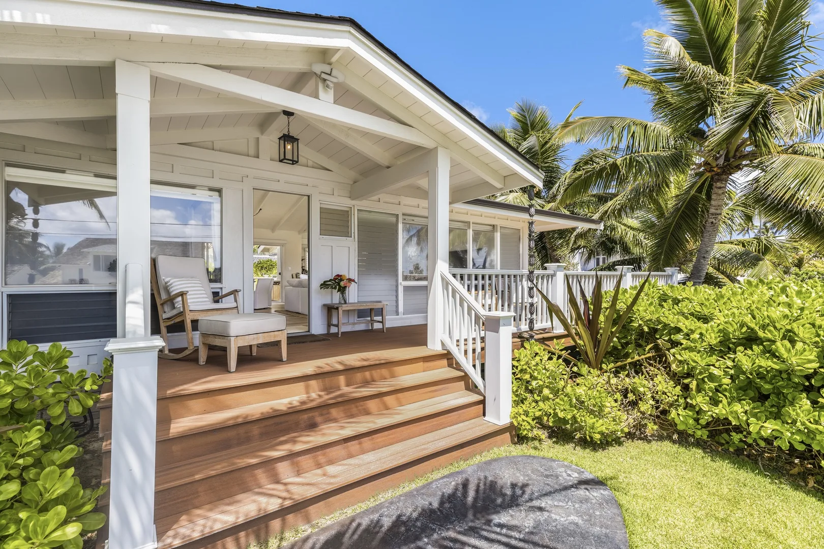 A white house porch with wooden steps, seating, and tropical plants.