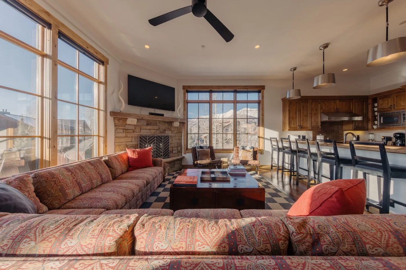 A living room with patterned seating, a stone fireplace, and an open kitchen.