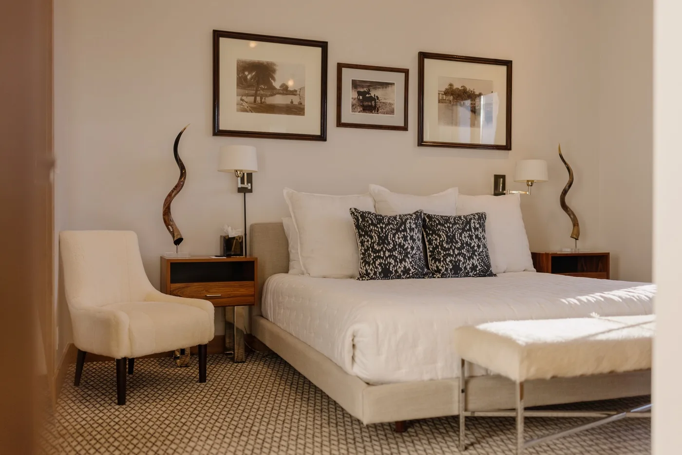 A bedroom with a neatly made bed, framed pictures, and a cushioned seat.