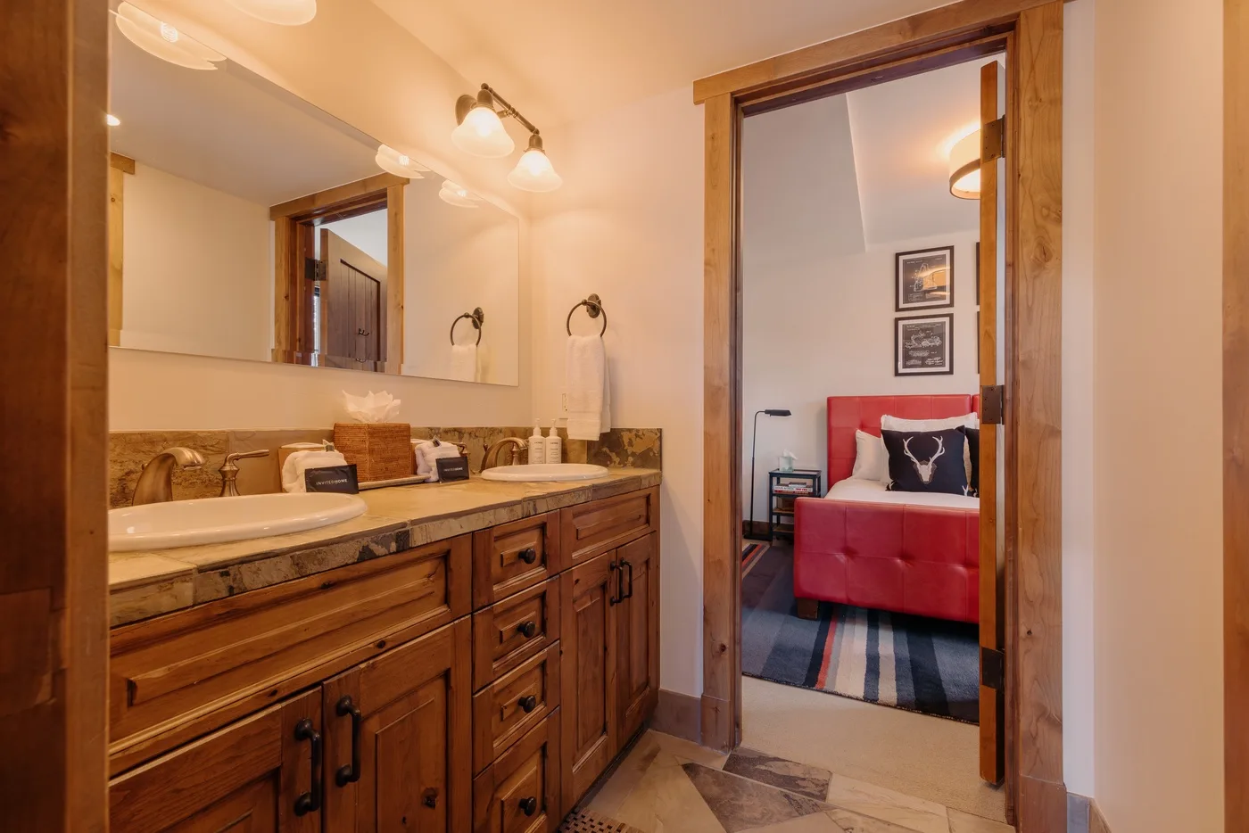 A bathroom vanity with a wide mirror opens onto a bedroom with a red bed.