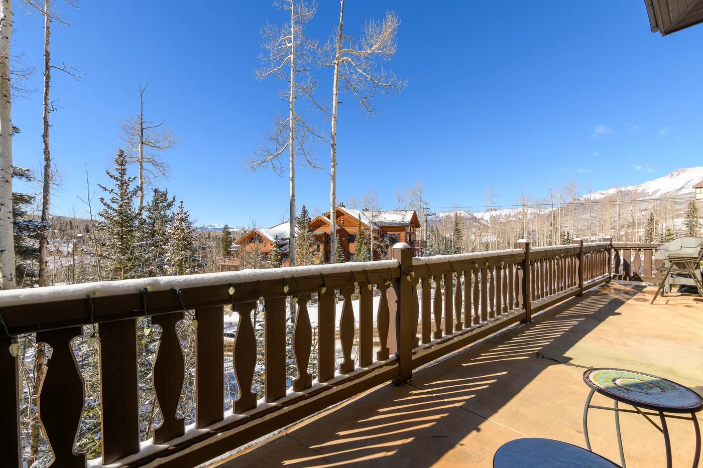A wooden deck overlooks trees, cabins, and a snowy mountain beneath a clear sky.