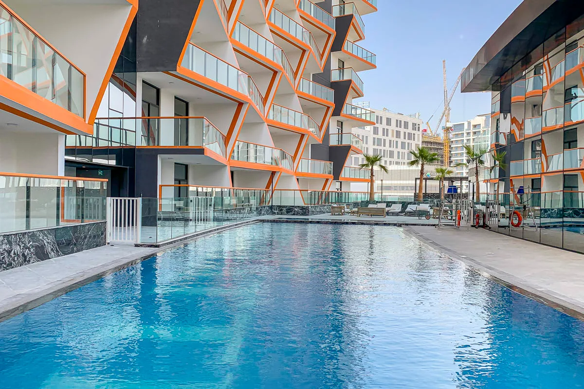 An outdoor swimming pool sits between modern buildings with angular orange balconies.