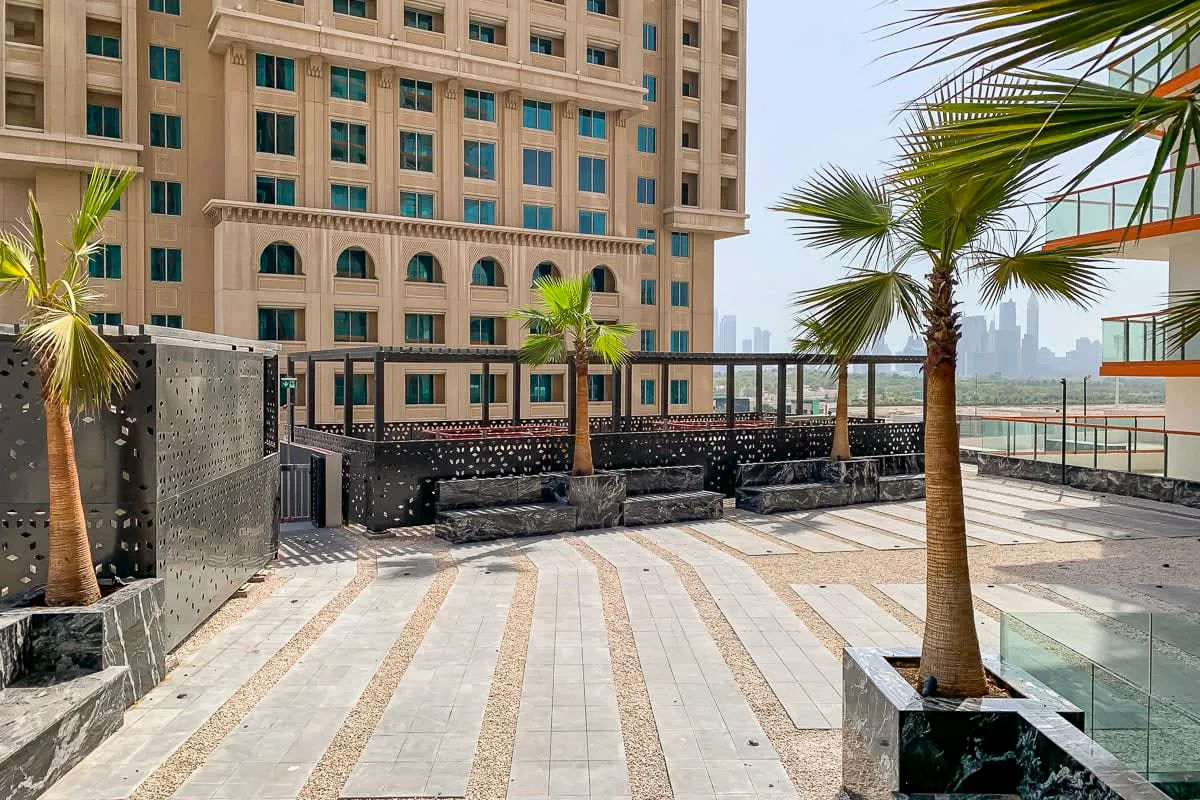 A paved outdoor terrace beside a high-rise building, with palm trees and seating.