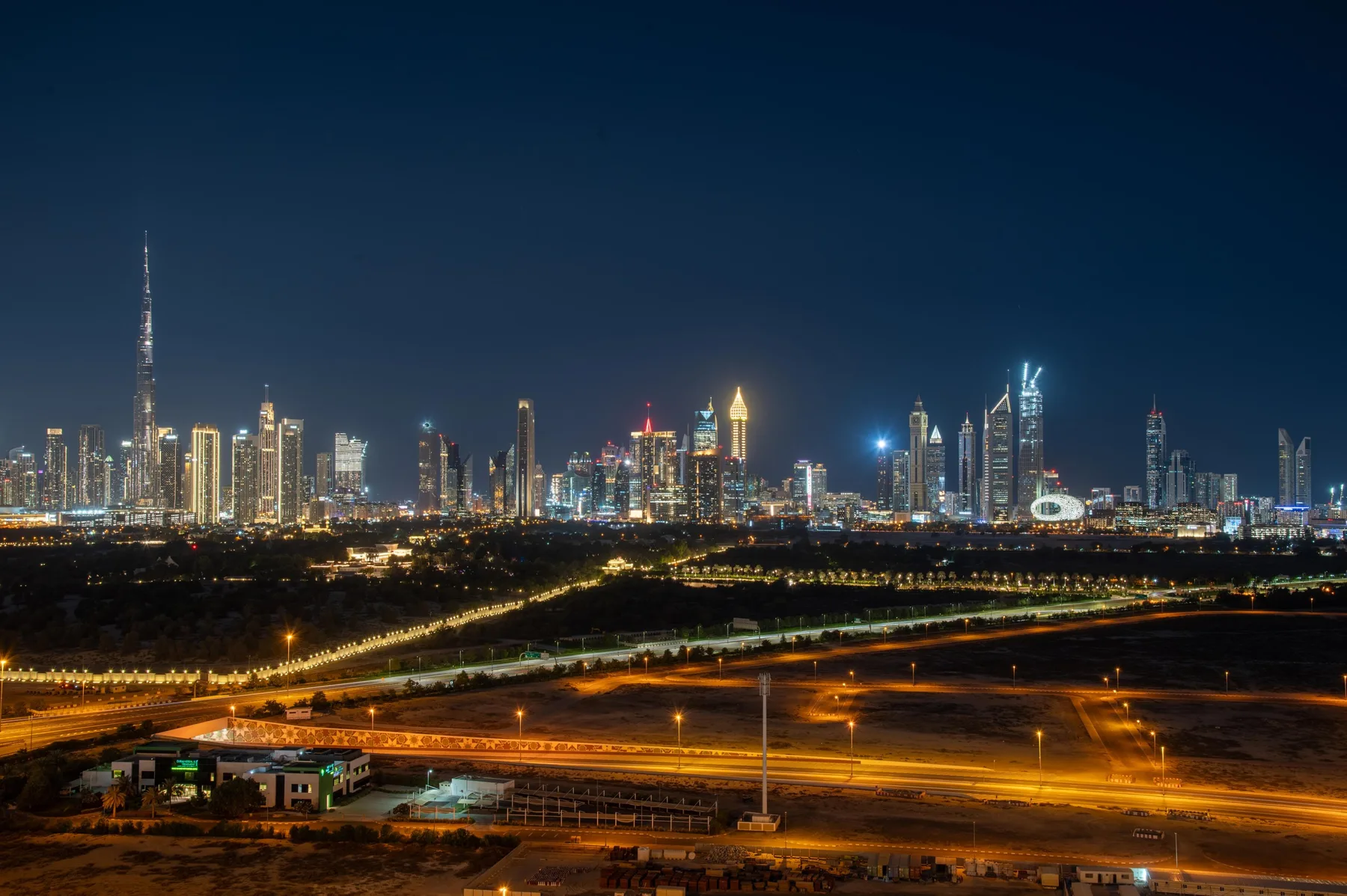 A nighttime city skyline glows beyond illuminated roads and open land.