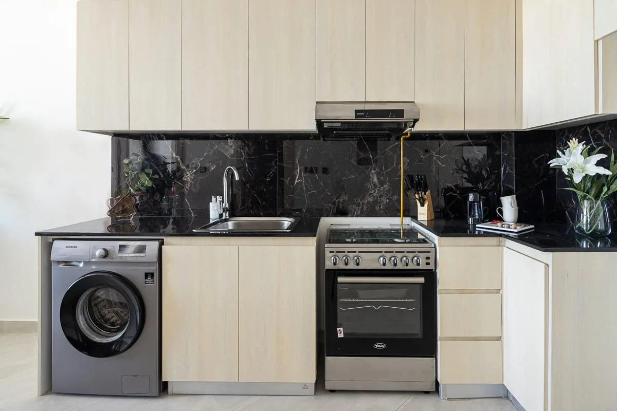 A kitchen with a stove, sink, and washing machine beneath pale cabinets.