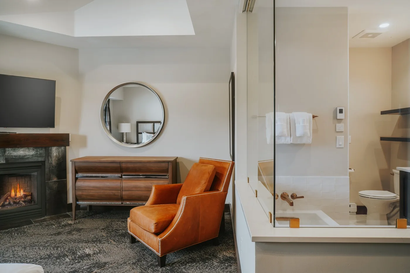 A bedroom sitting area with a dresser, round mirror, and fireplace beside a glass shower.