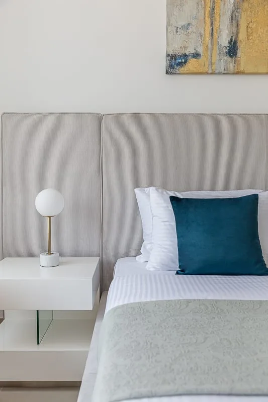 A neatly made bed with a teal pillow sits beside a small bedside table and lamp.