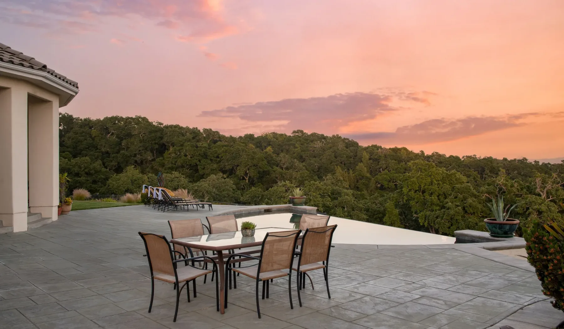 Una terraza exterior de baldosas tiene una mesa de comedor y asientos frente a un paisaje boscoso al atardecer.