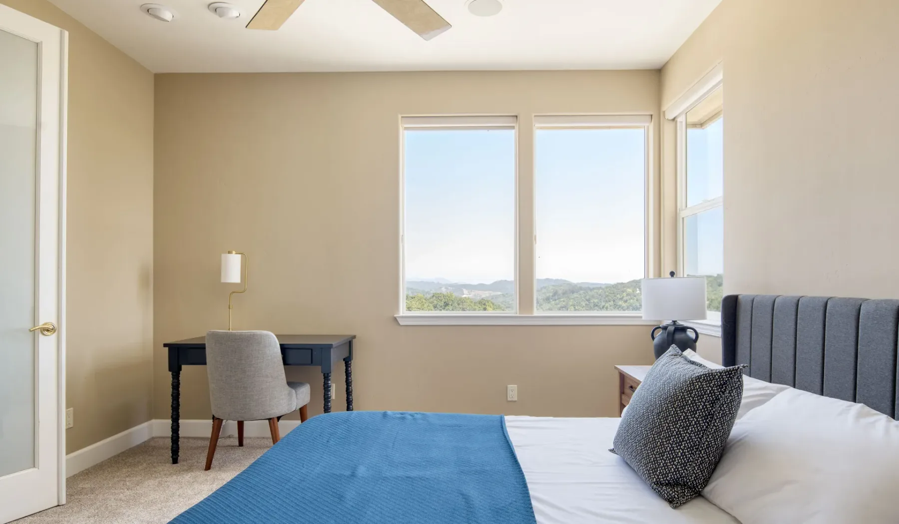 Un dormitorio con una cama, una colcha azul y un escritorio junto a las ventanas.