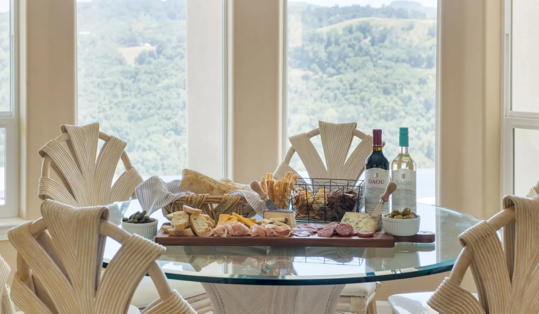Una mesa de comedor está dispuesta con aperitivos y bebidas junto a grandes ventanas con vistas a colinas verdes.