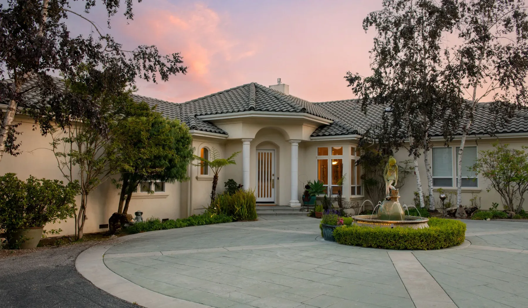 Una casa grande con entrada curva y fuente ajardinada al anochecer.