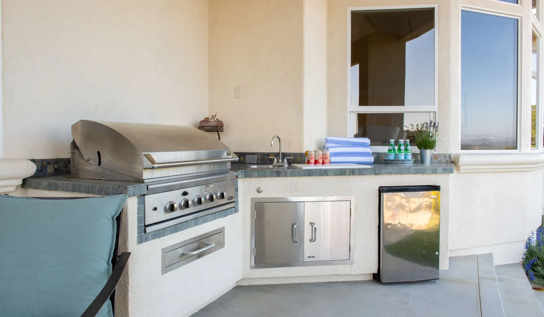 Una cocina al aire libre con una parrilla de acero inoxidable y una encimera bajo grandes ventanas.