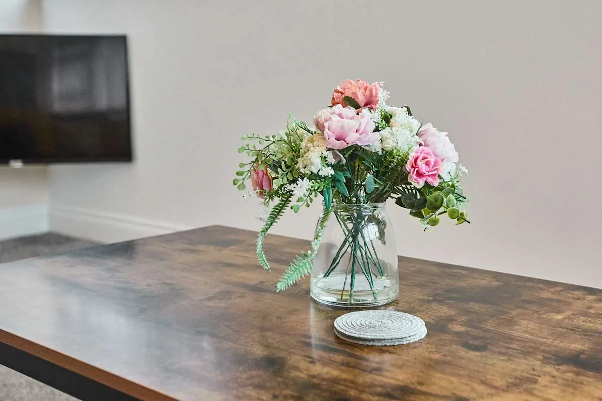 Un arreglo floral en un jarrón de vidrio está sobre una mesa, debajo de un televisor en la pared.